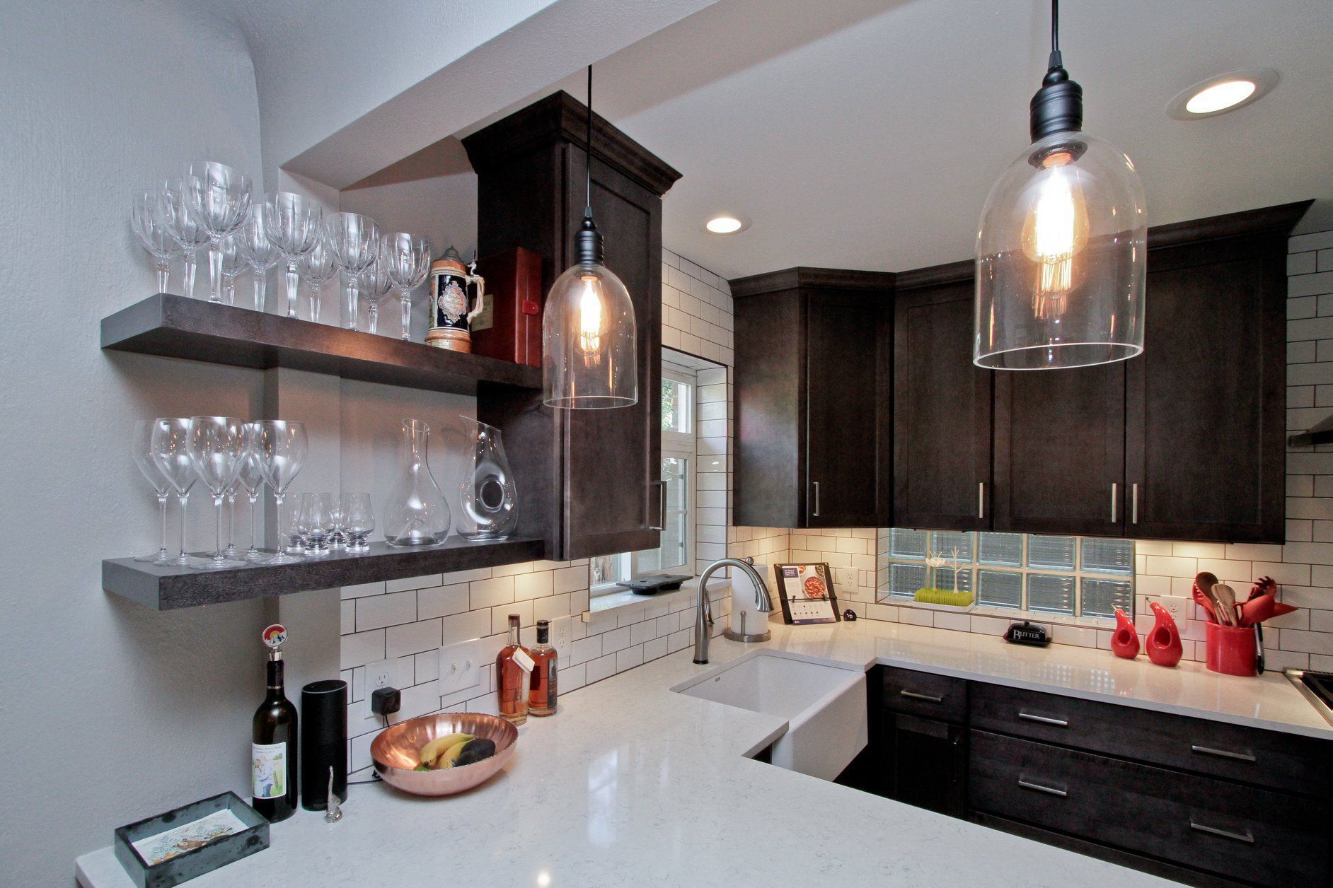 A kitchen with a sink , shelves , bottles and glasses on the counter.