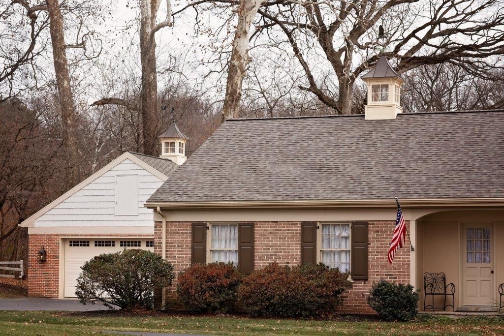 House with a garage, brick exterior, shutters, and an American flag. Trees in the background.