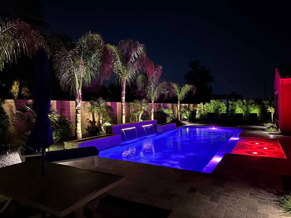 A large swimming pool is lit up at night with palm trees in the background.