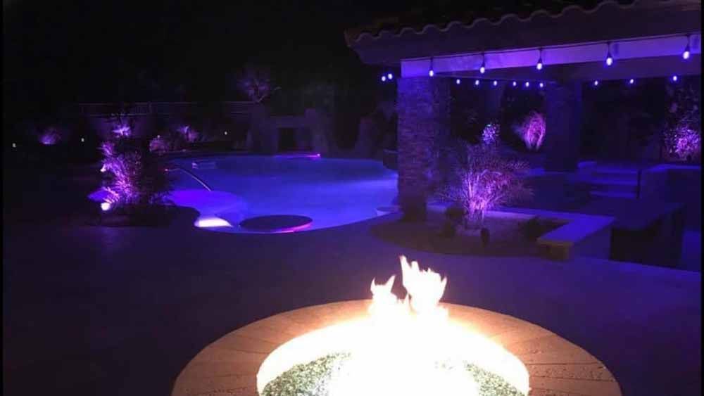 A fire pit is lit up at night in front of a pool.