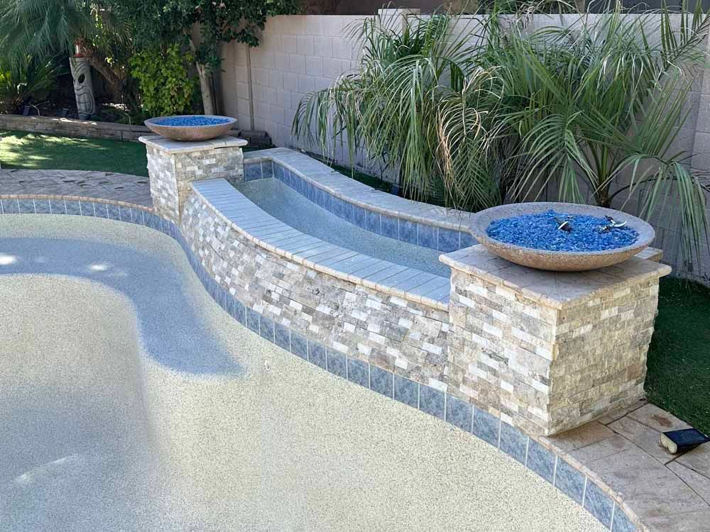 A swimming pool with a stone wall and two bowls of fire glass on the edge.