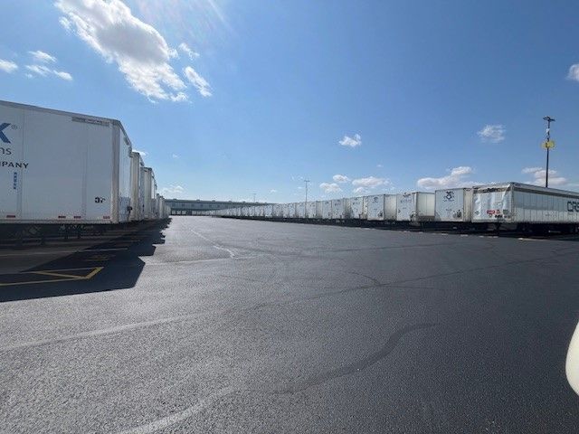 Two rows of white semi-trailers parked in a vast, empty asphalt lot under a sunny blue sky. 