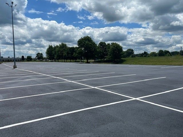 An empty asphalt parking lot with painted white parking lines under a partly cloudy blue sky, bordered by trees and grass.