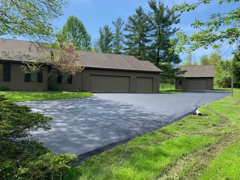 A brown house and detached garage with a newly paved asphalt driveway surrounded by green lawn and trees.