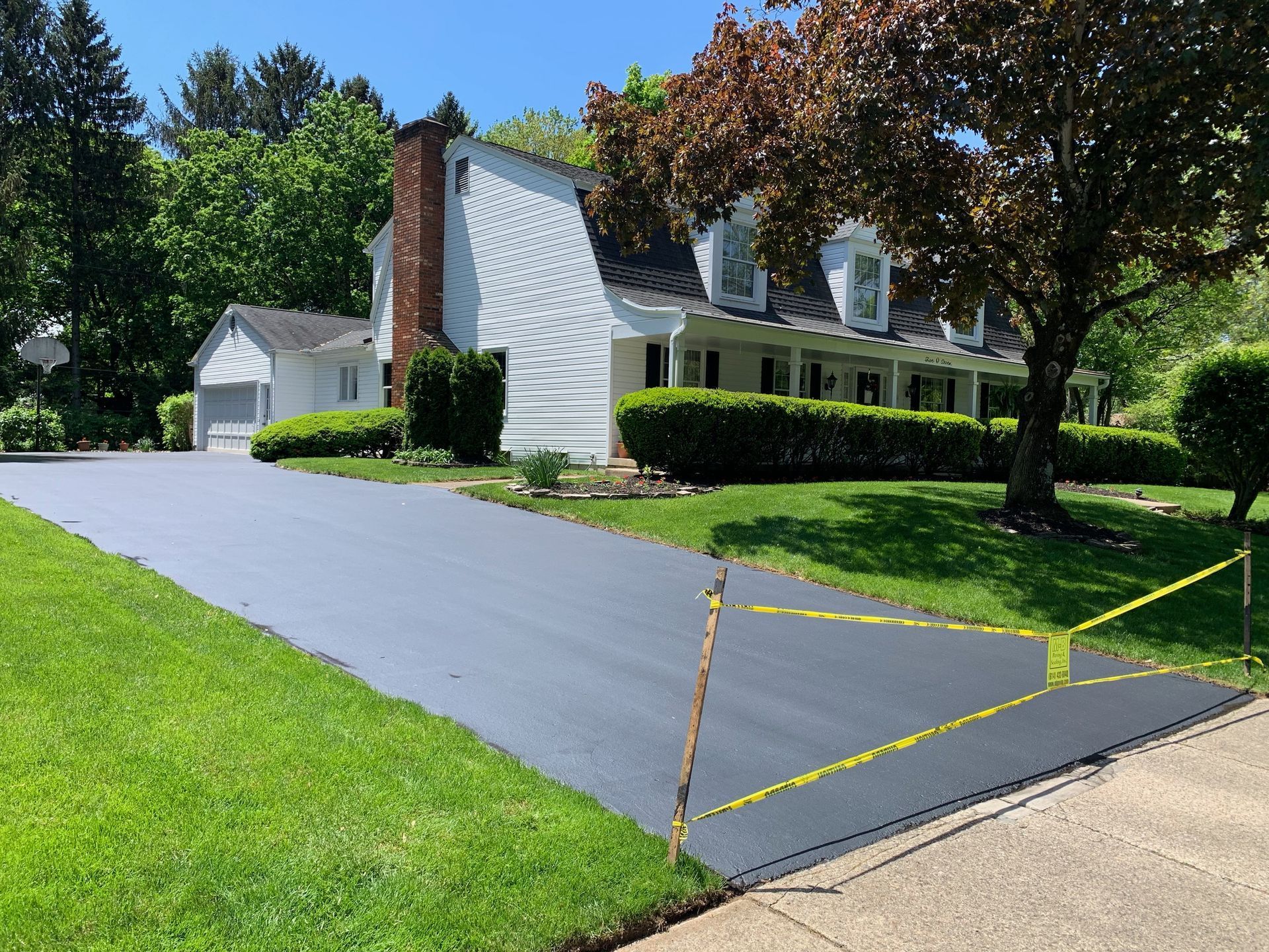 A freshly paved, smooth dark gray driveway leads up to a white house with a brick chimney and green lawn.