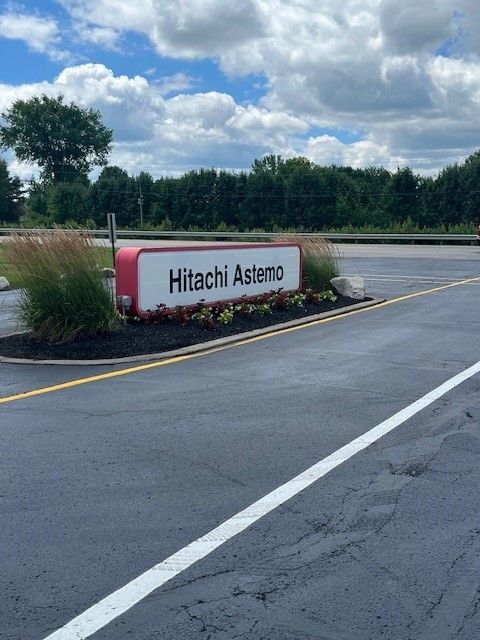 A red and white Hitachi Astemo sign sits in a landscaped garden next to a paved road under a partly cloudy blue sky.