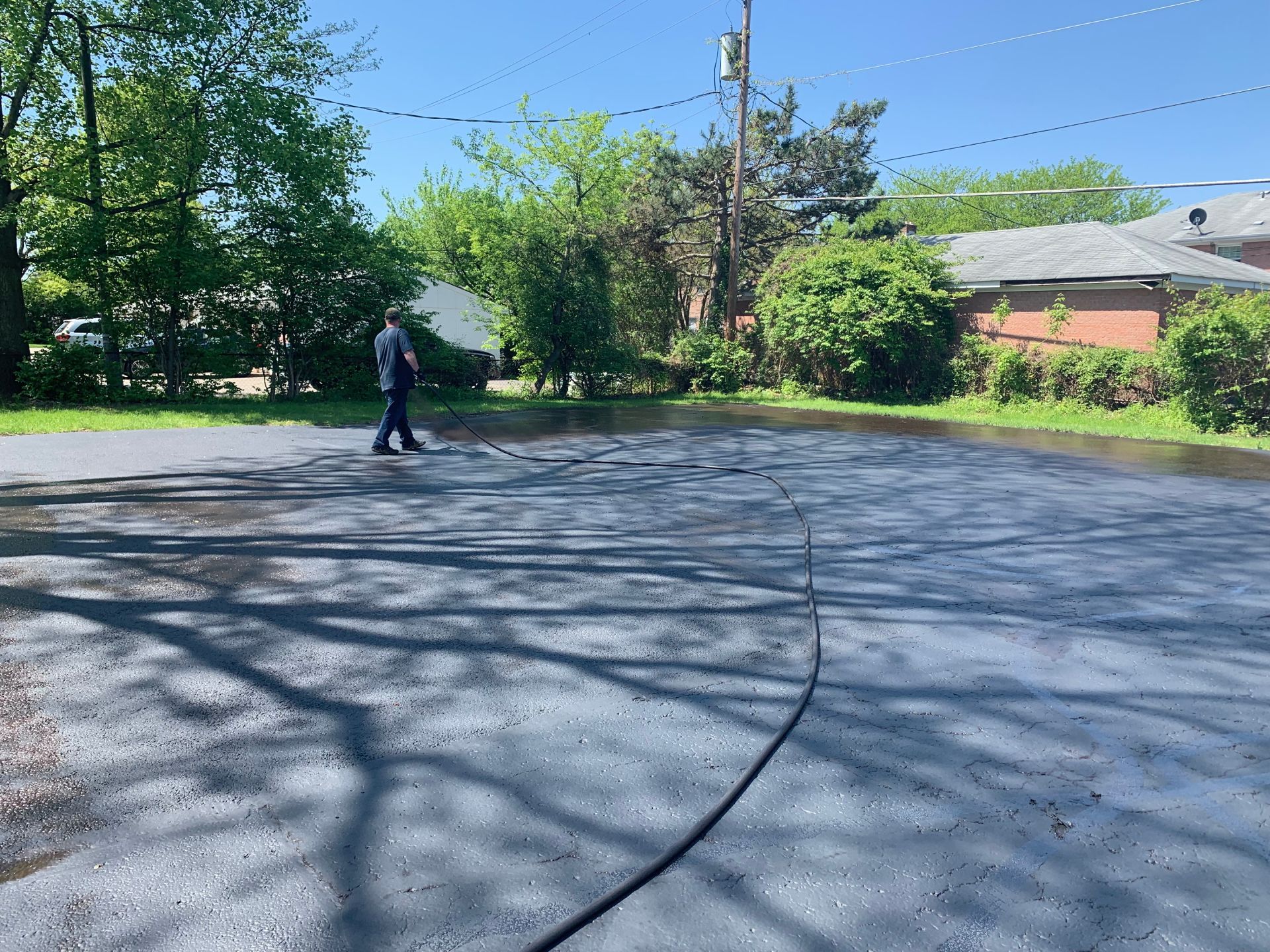 A person uses a long-handled tool to apply dark sealant to a large, paved residential driveway on a sunny day.