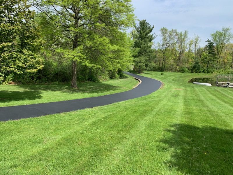 A black asphalt path curves through a lush green lawn, bordered by trees under a bright blue sky.