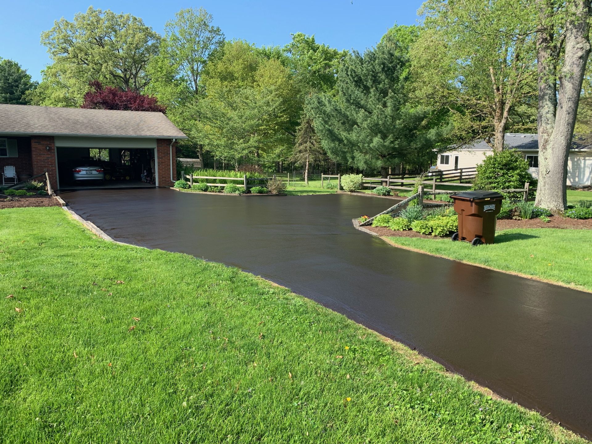 A freshly sealed, dark asphalt driveway leads to a brick garage at a home surrounded by lush green lawns and trees.