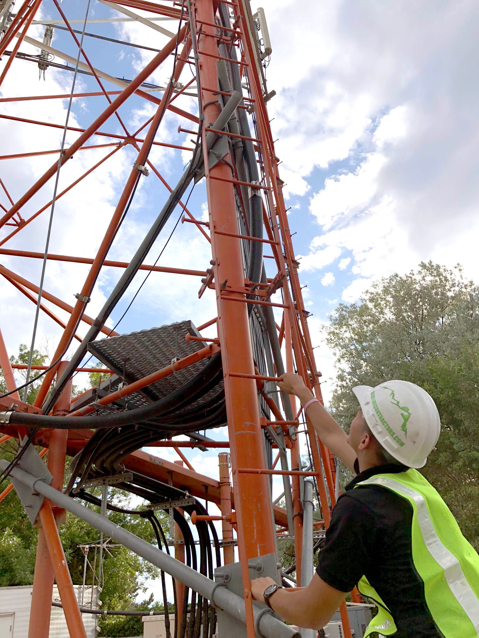Person in safety vest inspecting a tall orange communication tower with cables. Cloudy sky visible.