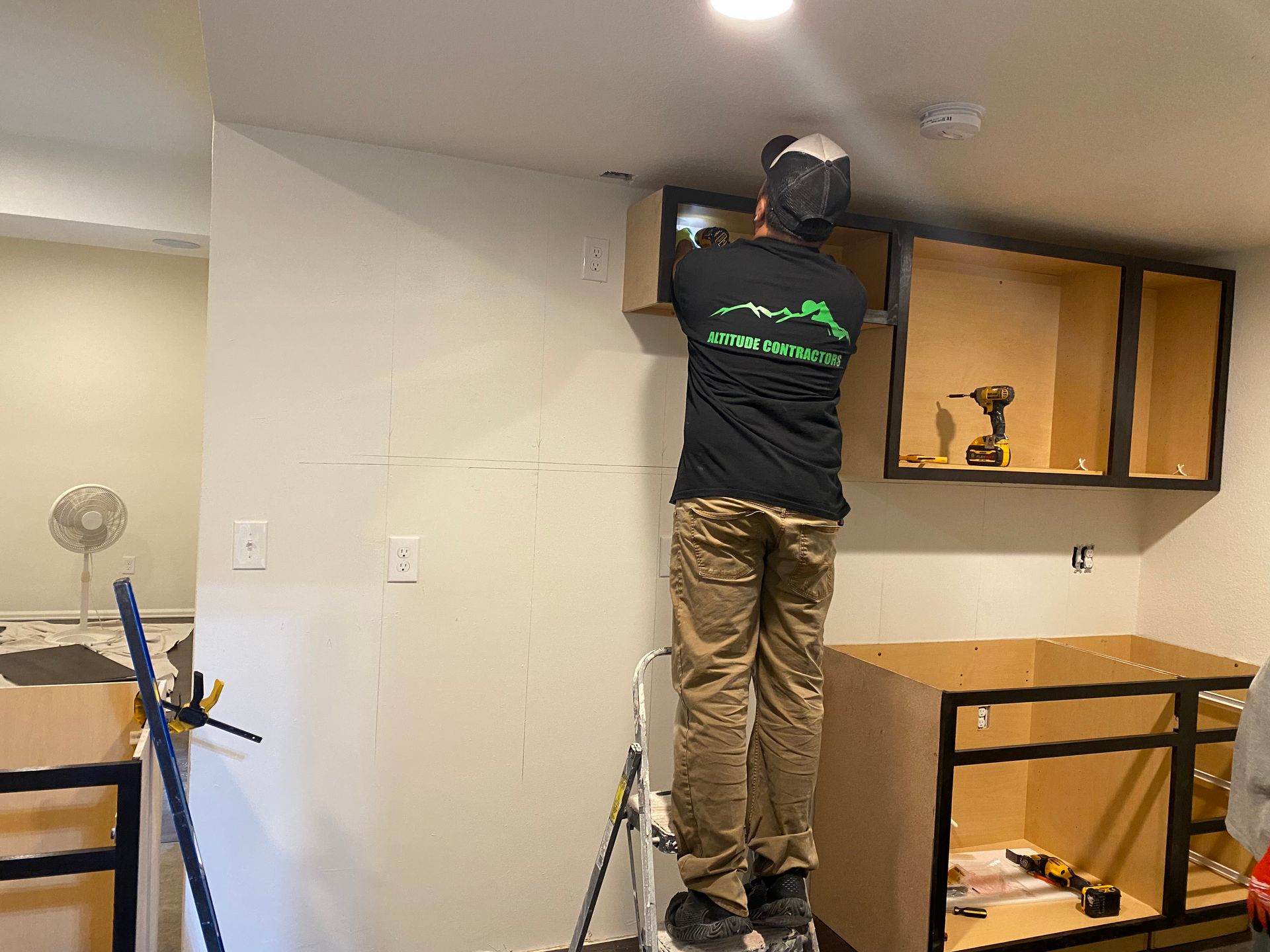 A person on a ladder installs a cabinet in a kitchen, using tools.