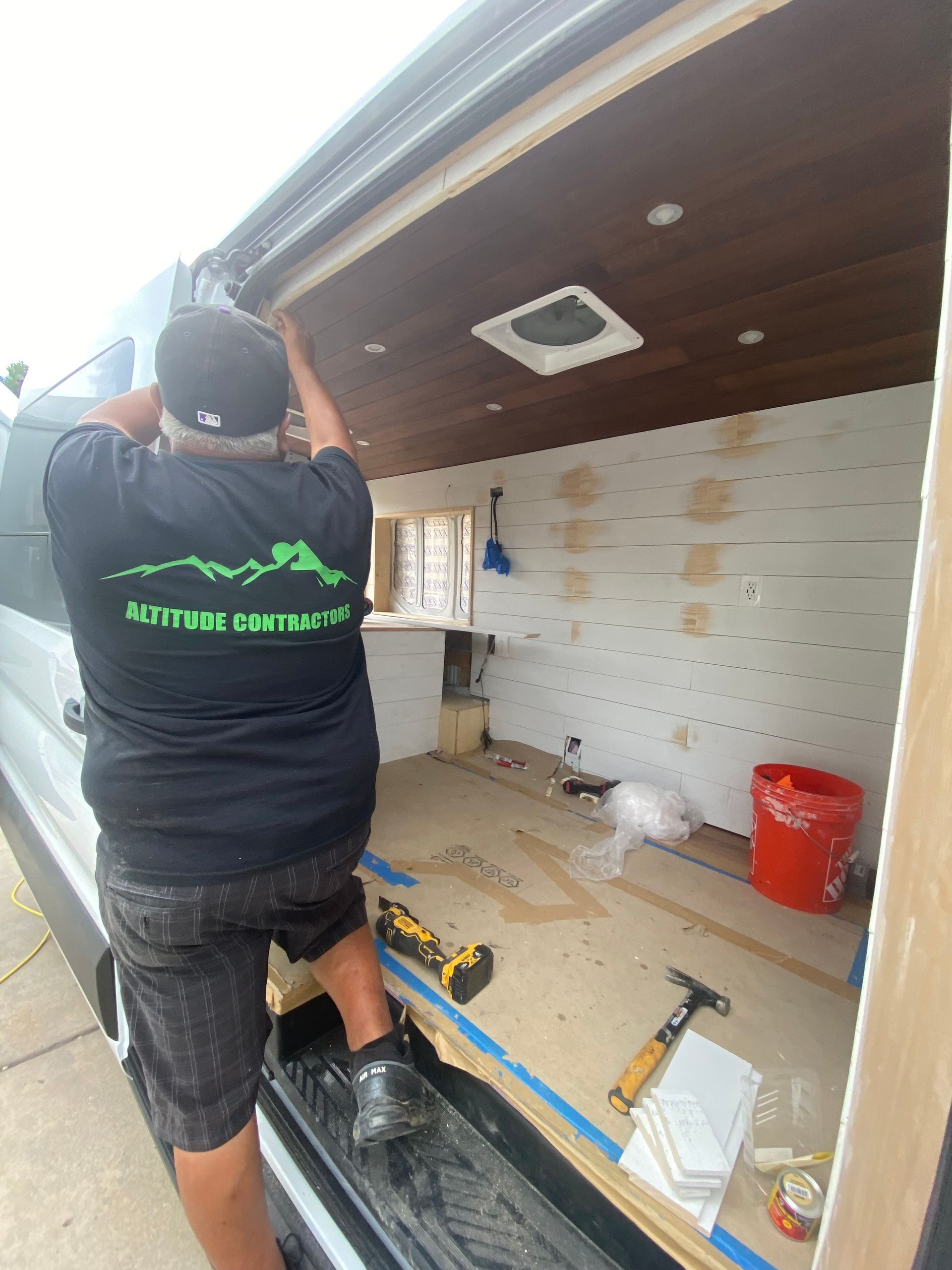 Person installing trim inside a van, wood panel ceiling, white walls, tools and materials present.