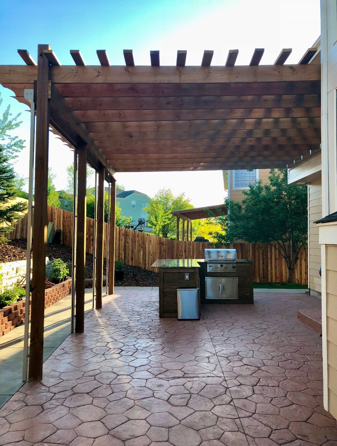 Wooden pergola covers a stamped concrete patio with an outdoor kitchen, looking toward a fence and trees.
