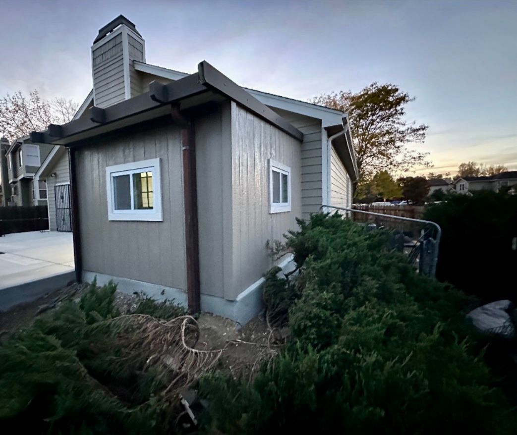 Tan-sided building with two windows and a chimney, next to bushes under a cloudy sky.