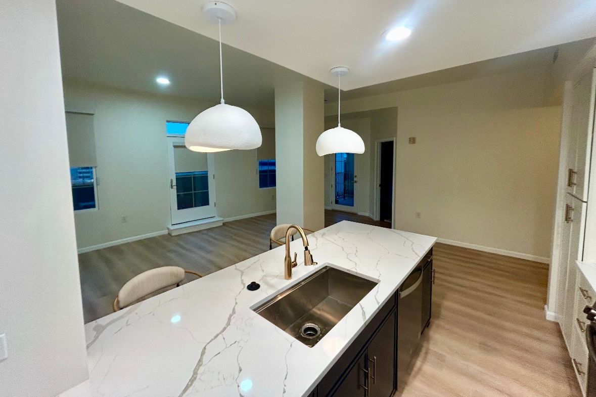 Modern kitchen with white countertops, sink, pendant lights, and view into living area.