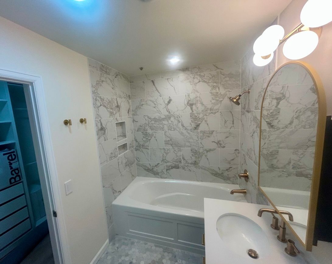Bathroom with marble tile, white tub, gold fixtures, and an arched mirror.