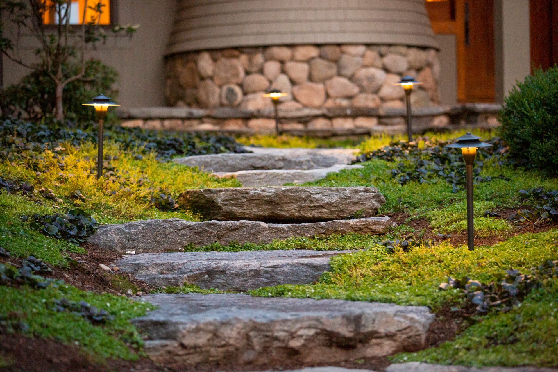 A stone walkway leading to a house with lights on the side of it.