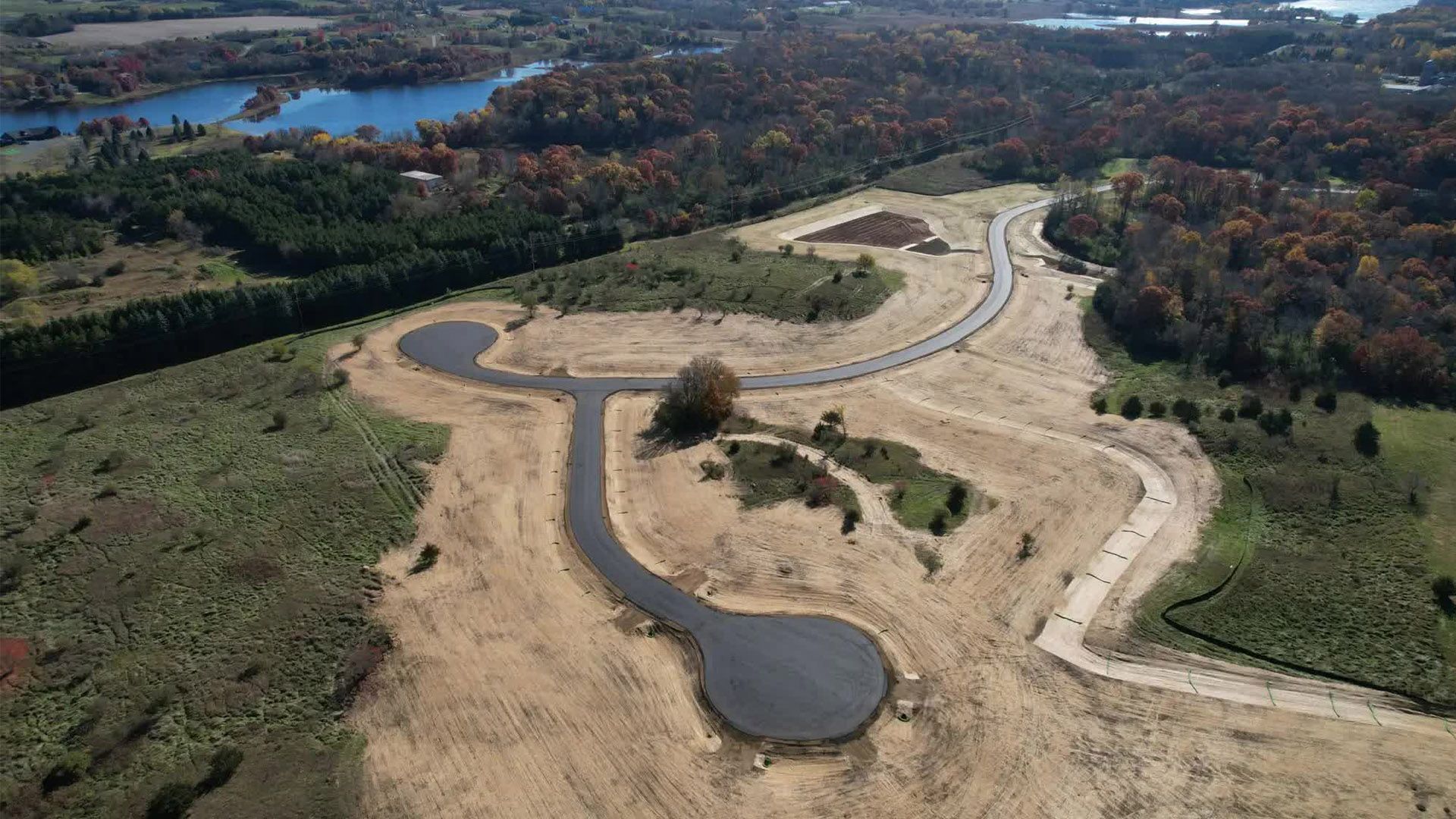 Aerial view of a new residential development with paved roads and cul-de-sacs surrounded by wooded areas and a lake.