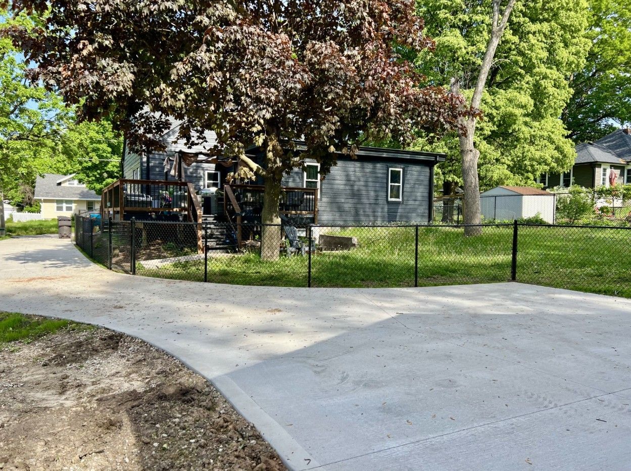 A grey house with a deck sits behind a black chain-link fence, viewed from a wide concrete driveway on a sunny day.