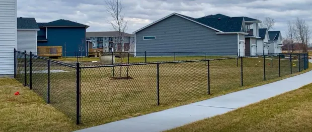 A chain link fence surrounds a grassy yard, sidewalk in foreground, houses in background under overcast sky.