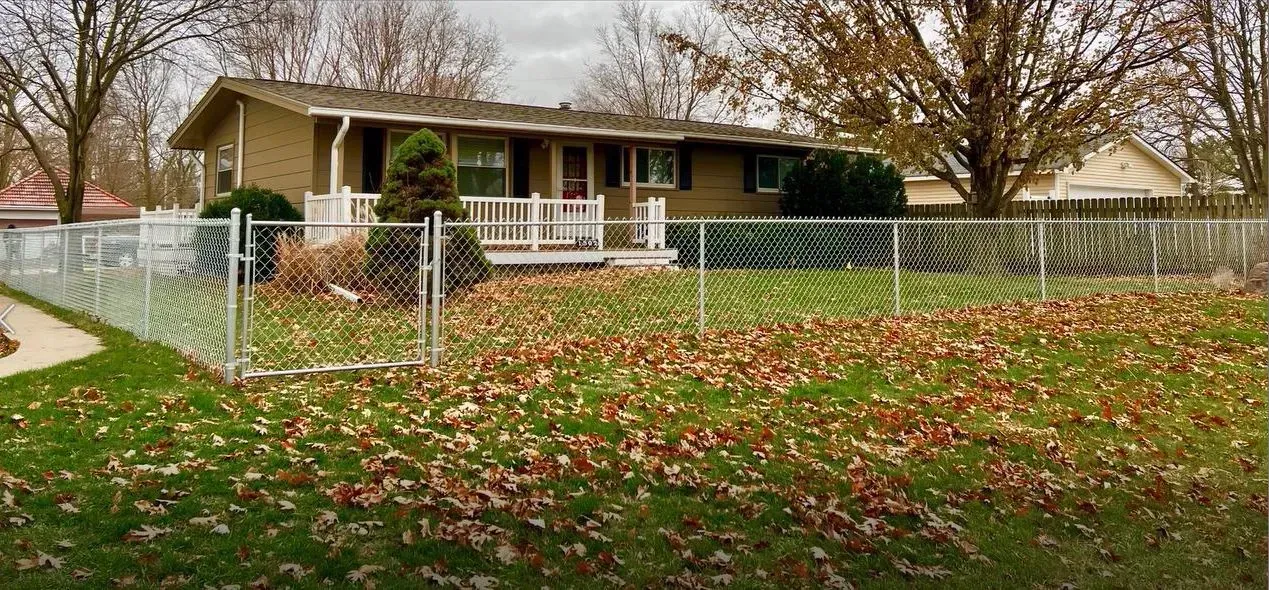 A house with a chain link fence in front; a yard with autumn leaves.
