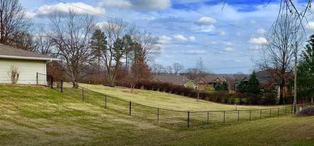 A grassy yard with a low fence, several leafless trees, and houses under a partly cloudy sky.