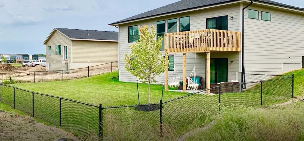 A house with a deck and lawn, enclosed by a black chain-link fence. Another house is in the background.