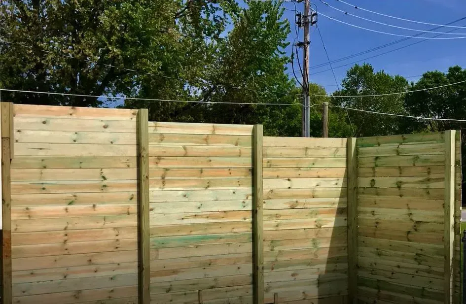 Wooden fence in front of green trees and a utility pole against a blue sky.