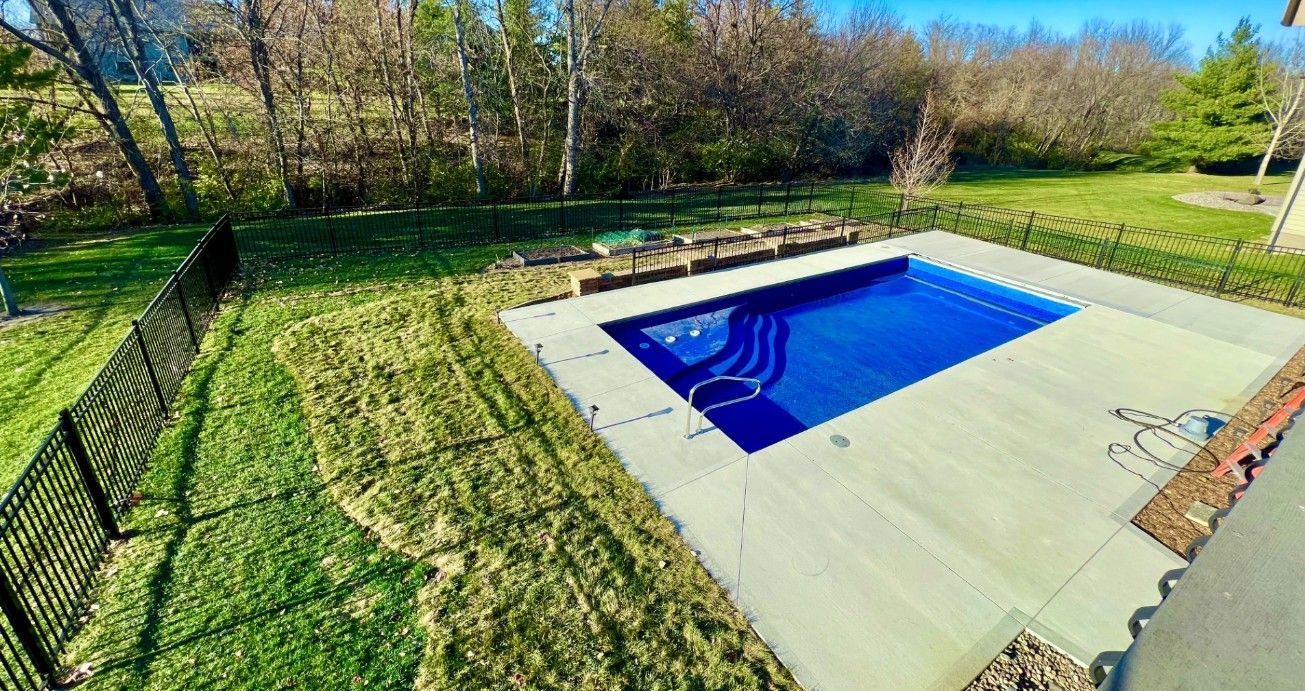 A rectangular blue swimming pool set in a backyard patio, surrounded by a black fence and a grassy lawn with trees.