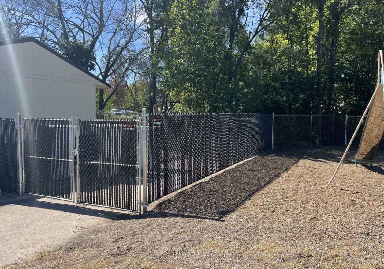 A black chain-link fence with privacy slats borders a mulched garden bed next to a building on a sunny day.
