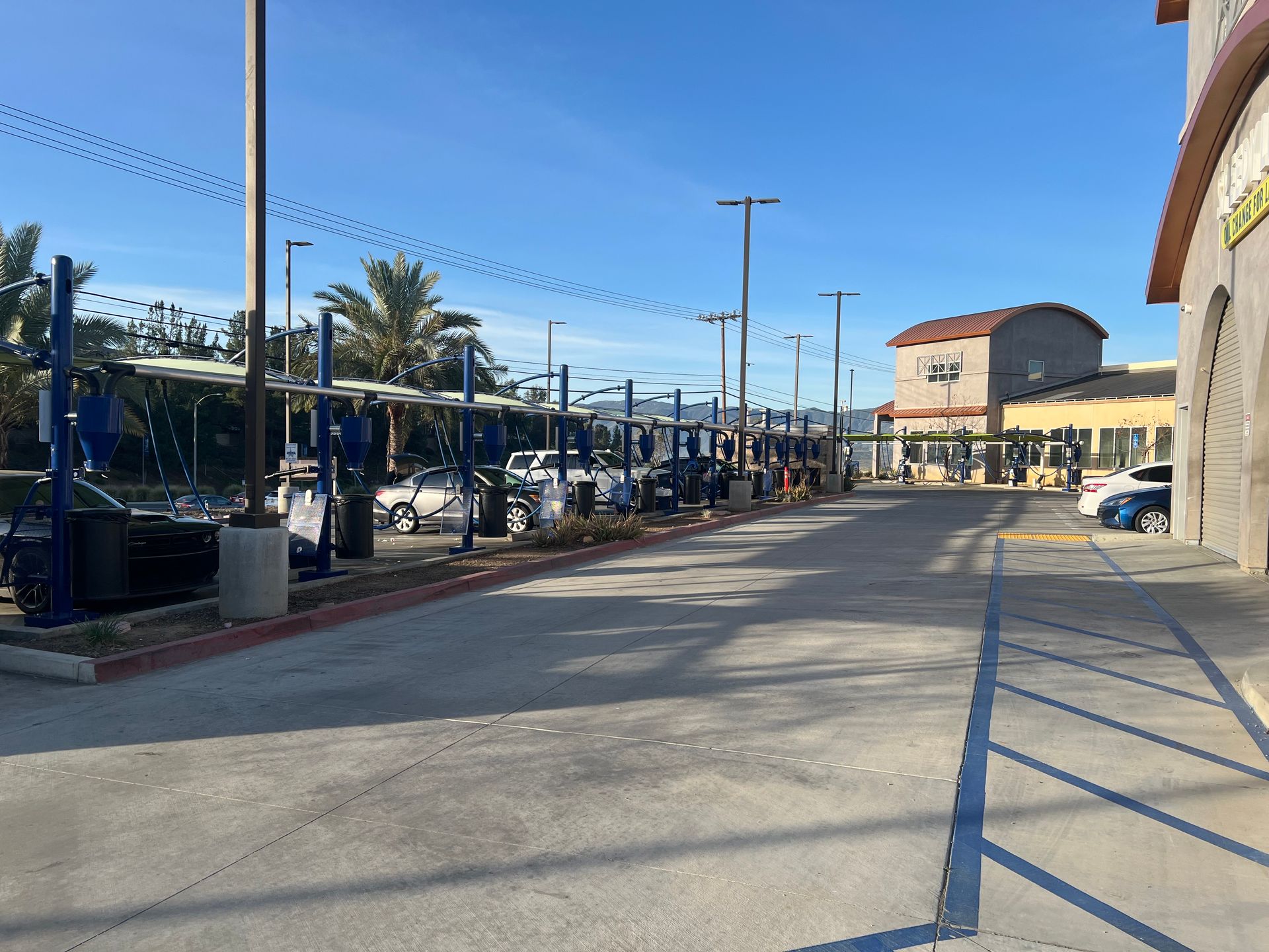 An outdoor car wash facility with blue support structures, parked vehicles, and a beige building under a clear blue sky.