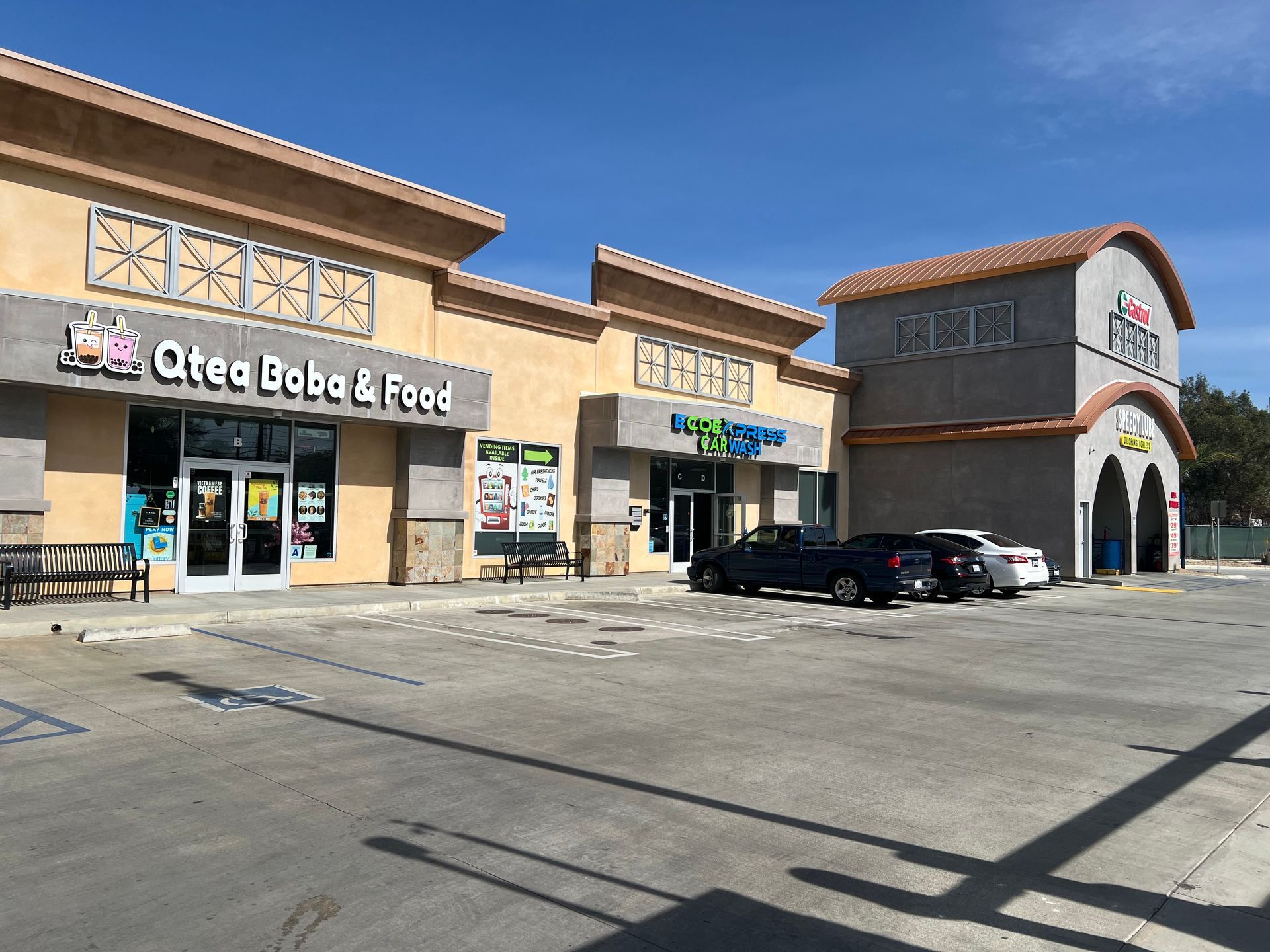 A single-story strip mall with a tan facade under a clear blue sky, featuring a Qtea Boba & Food shop and a parking lot.