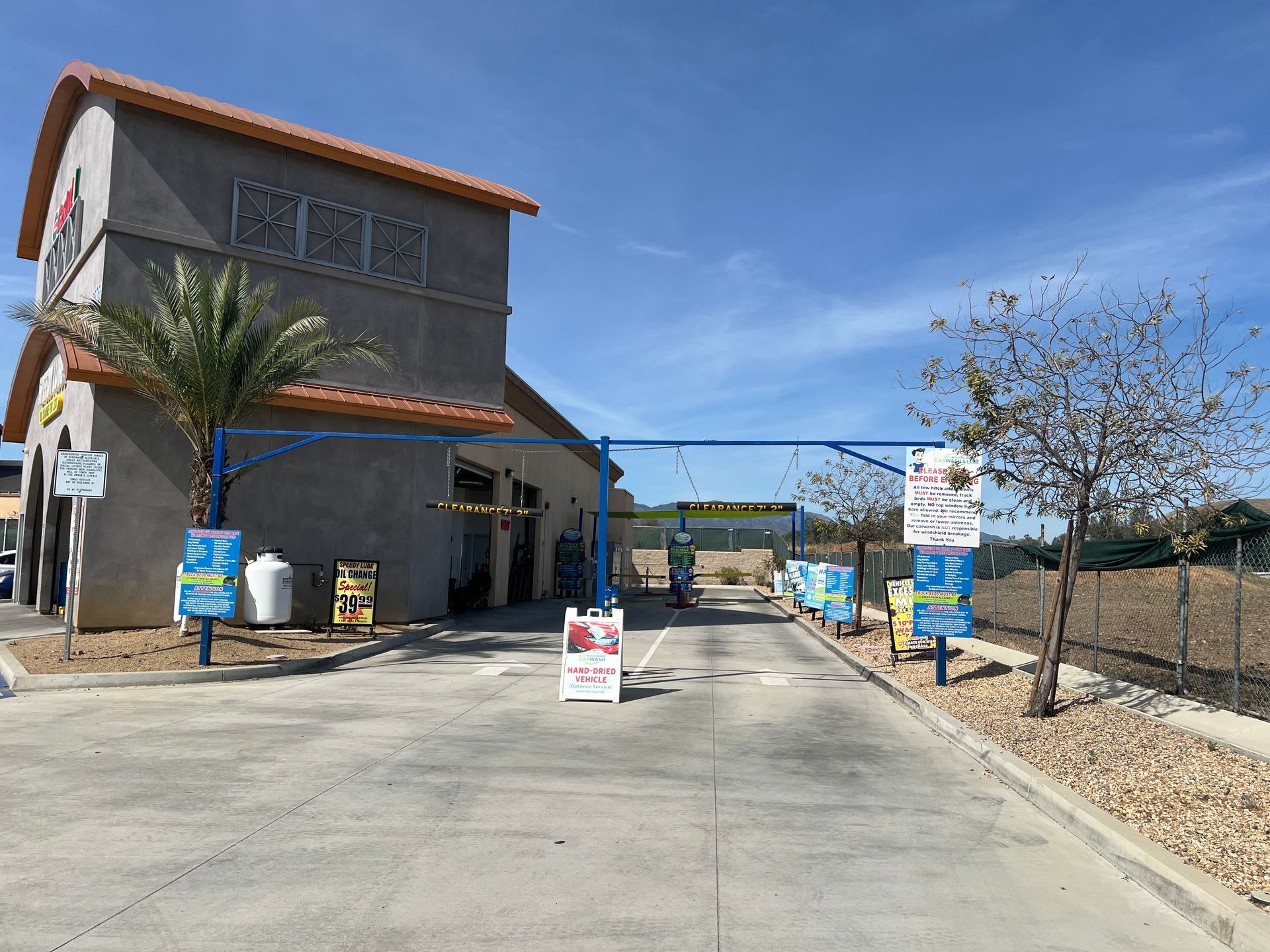 An outdoor car wash entrance with a beige building, a palm tree, blue metal arches, and informational signs under blue sky.