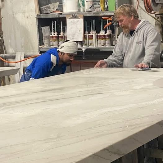 Two men examining a large, light-colored stone slab in a workshop, possibly for countertop fabrication.