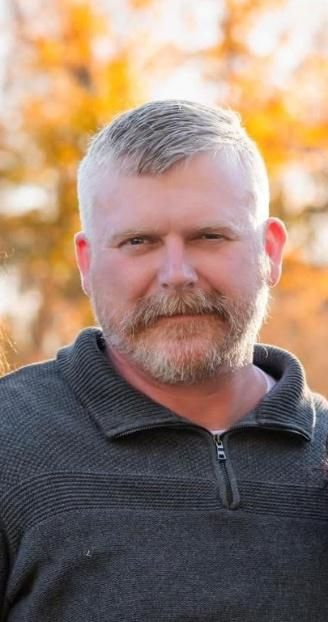 Man with short gray hair and a mustache, wearing a gray zip-up sweater, set against a blurred background of yellow foliage.
