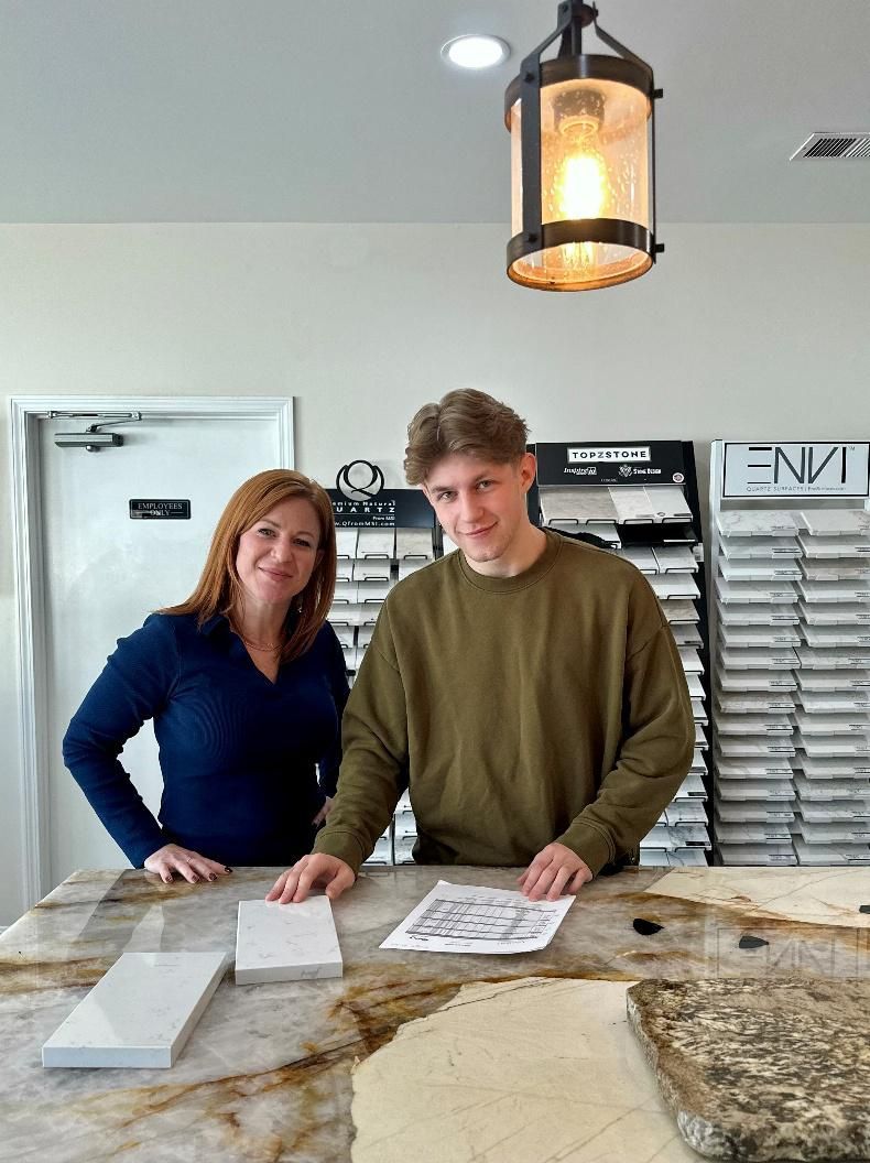 Woman and man at countertop, selecting tile samples and reviewing plans. Interior, lit by overhead lamp.