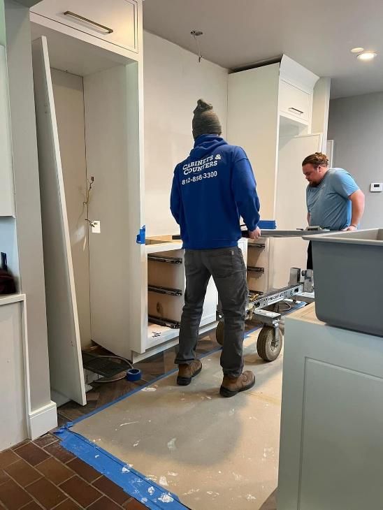 Two people moving a white kitchen cabinet on a wheeled cart in a kitchen with renovation in progress.