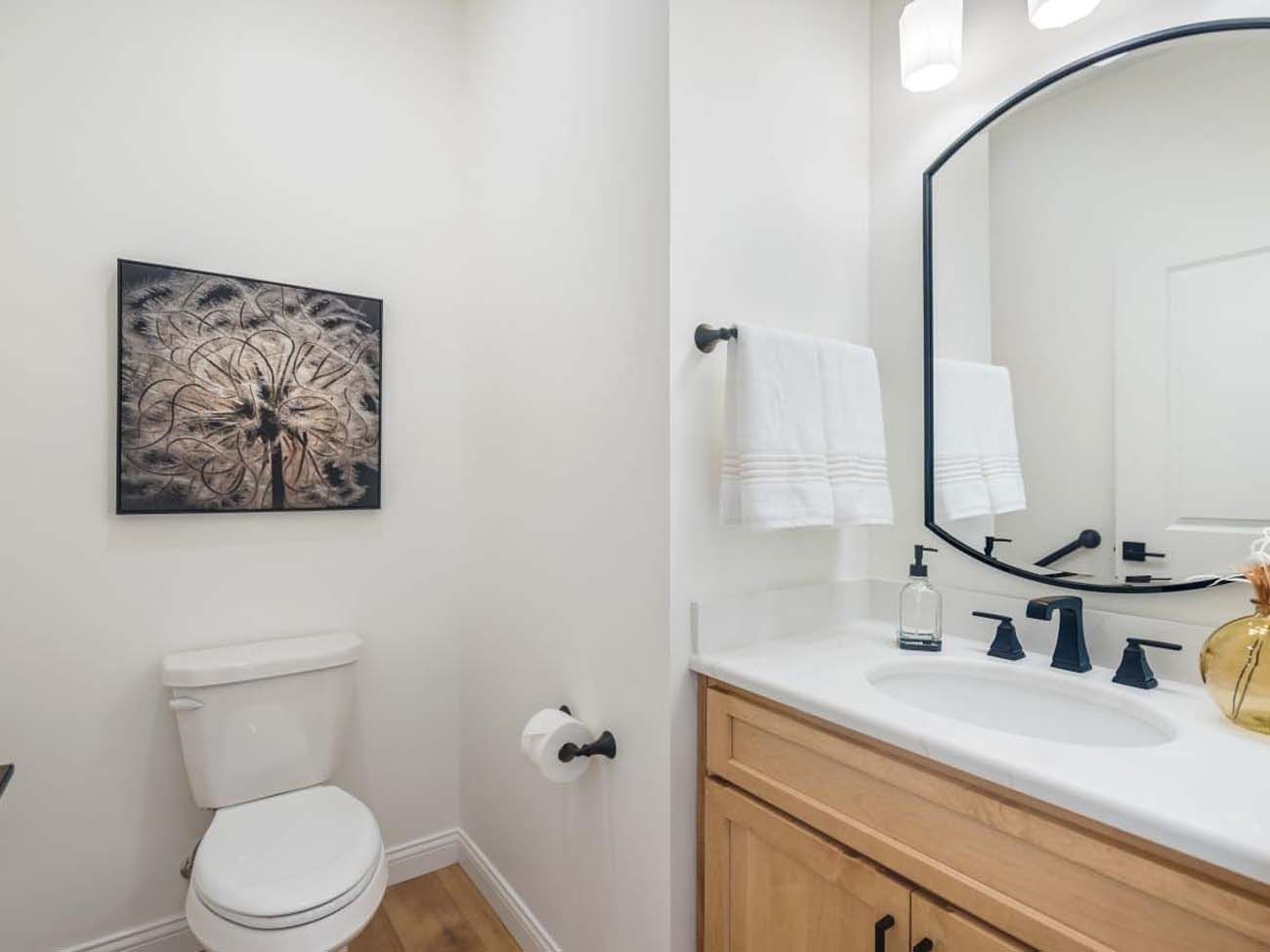 Bathroom with white walls, wooden vanity, round mirror, toilet, and artwork.
