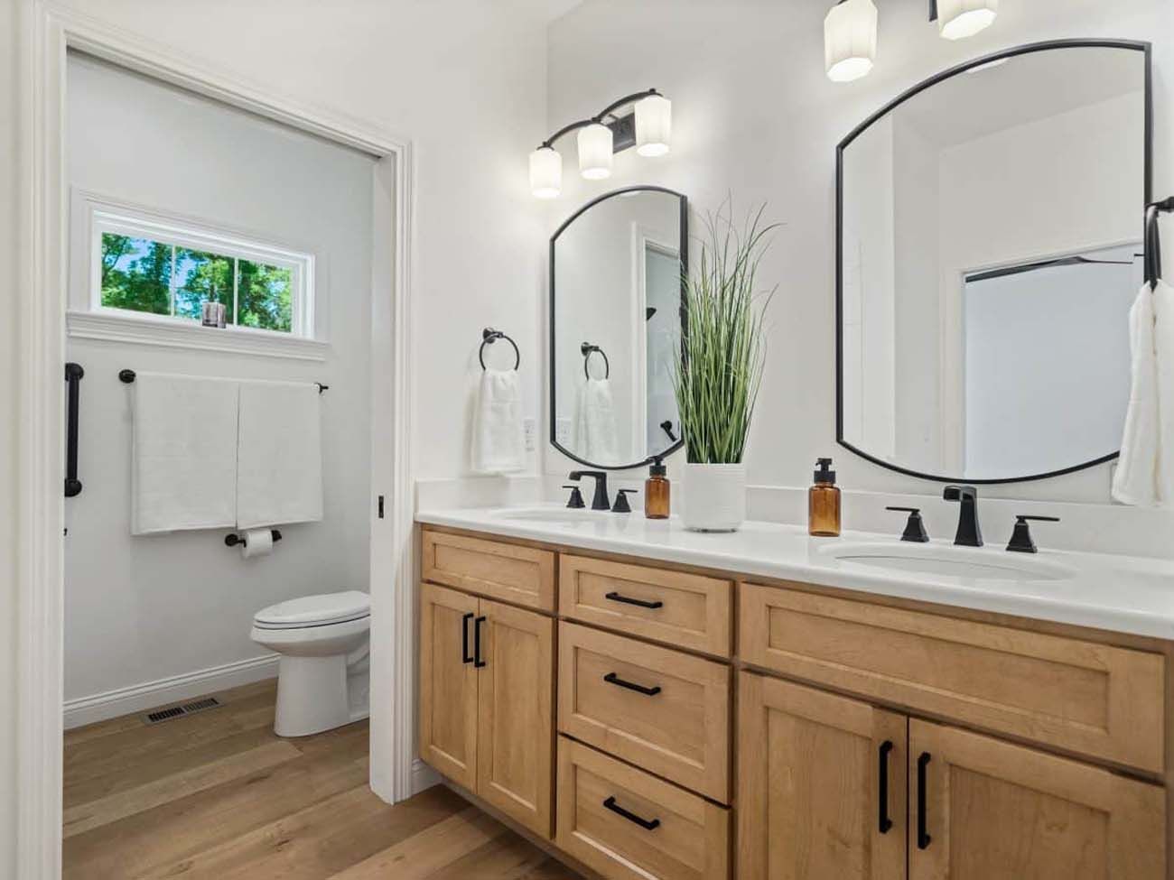 Bathroom with double vanity, two arched mirrors, wood cabinets, and toilet.