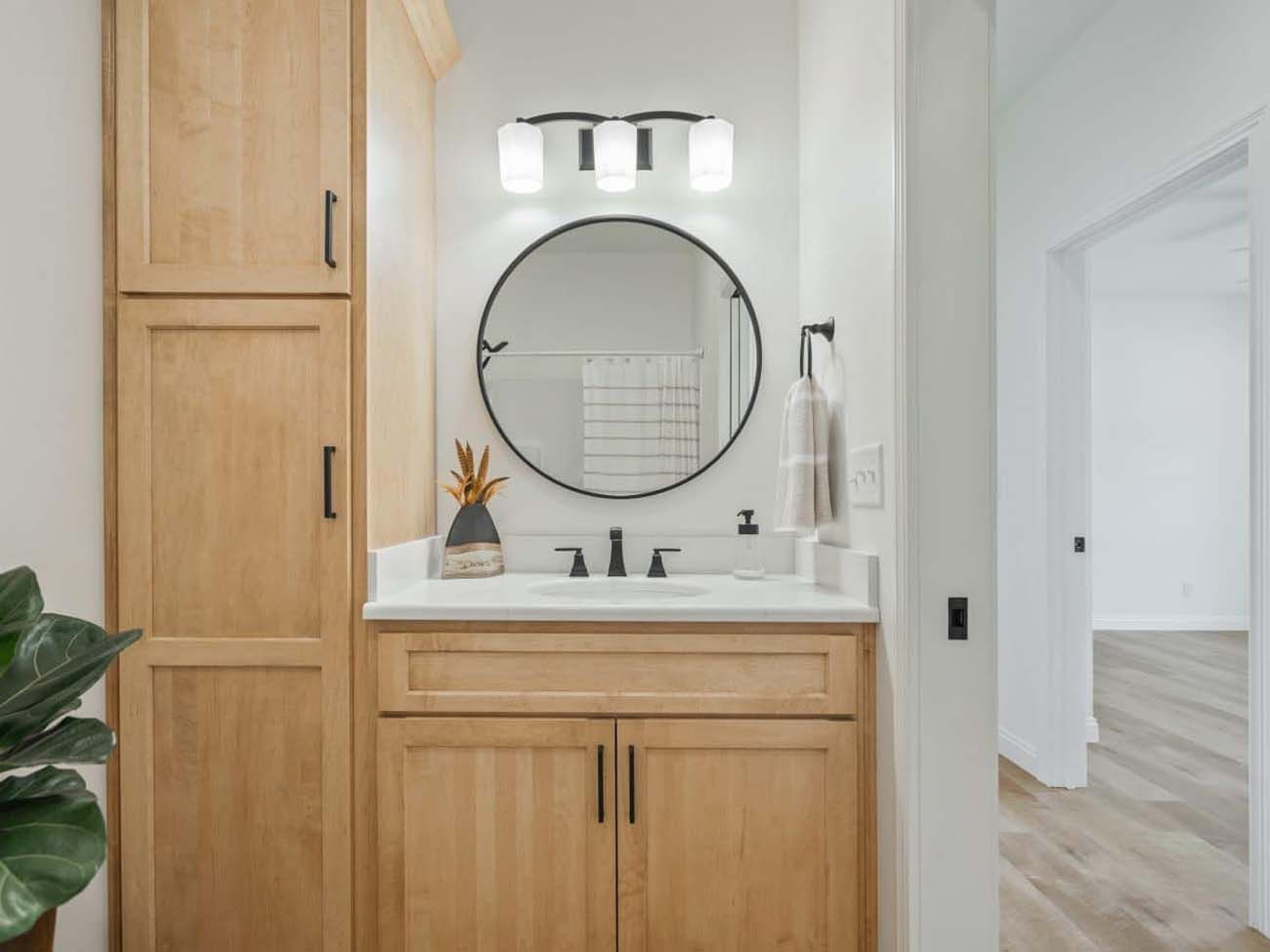 Bathroom with light wood vanity, round mirror, and tall cabinet. White walls and neutral decor.
