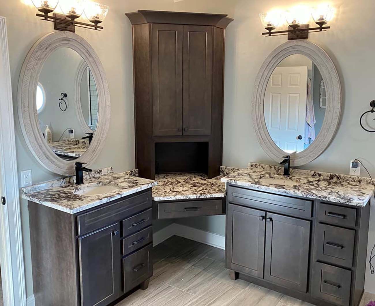 Bathroom with dual vanities, central storage cabinet, and oval mirrors. Dark wood cabinets with granite countertops.