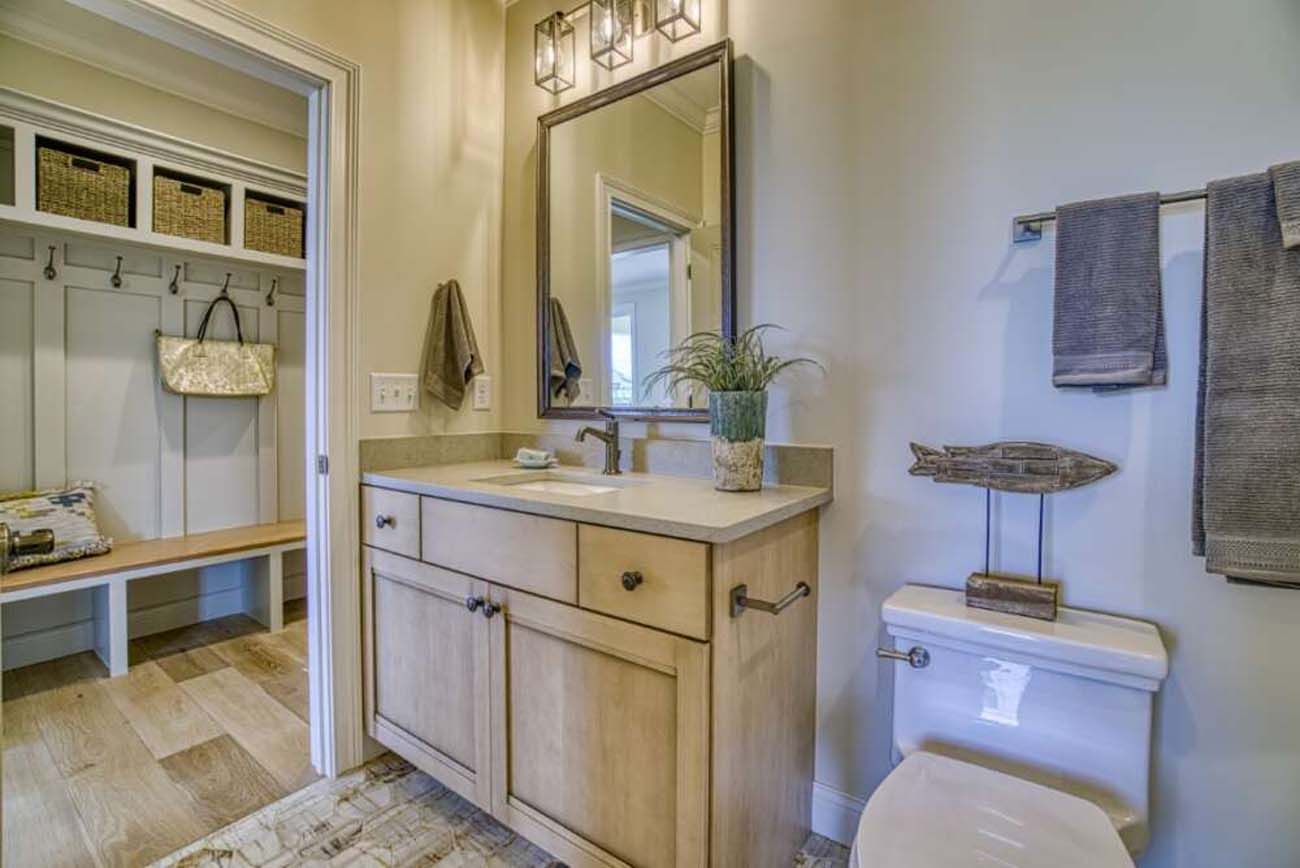 Bathroom with light wood vanity, mirror, and toilet. Open doorway to a hallway with a bench and storage.