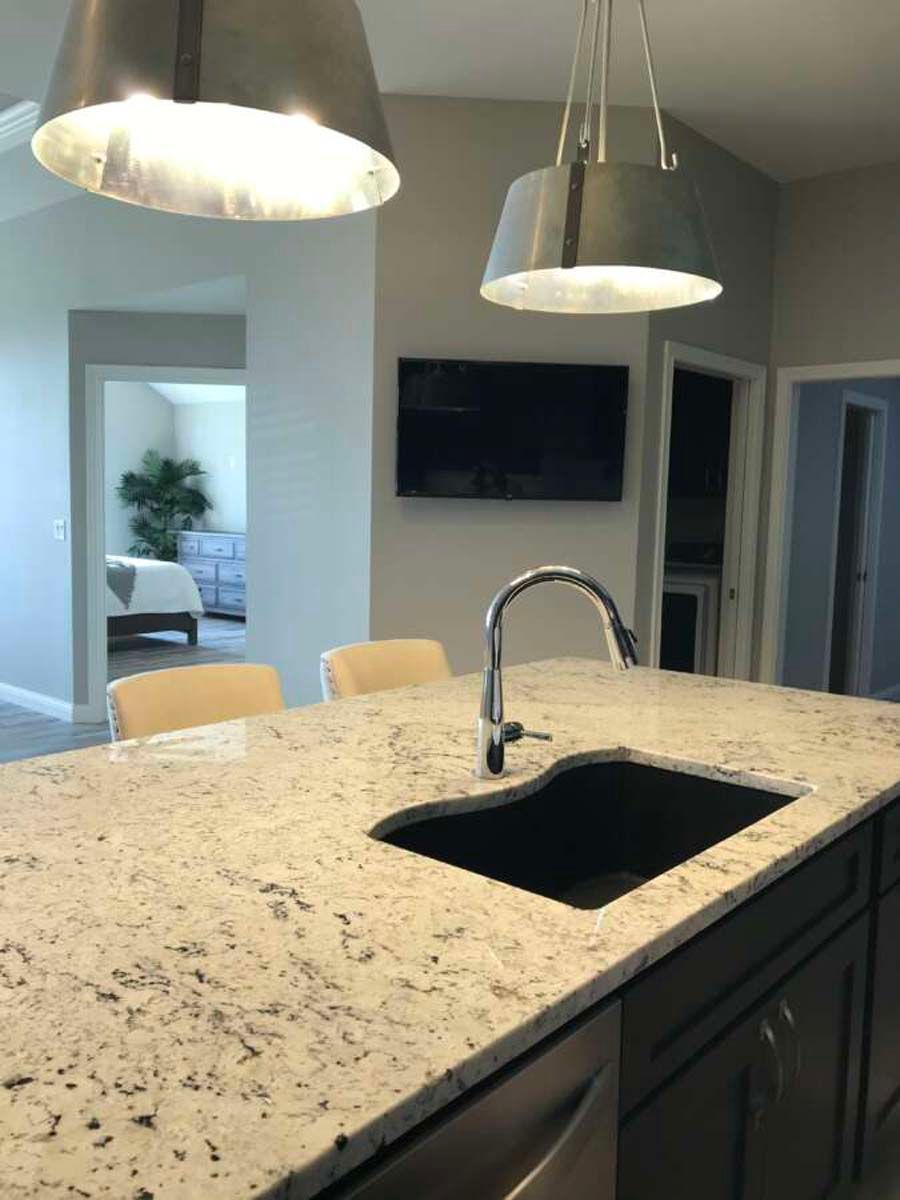 Kitchen island with granite countertop, sink, and overhead pendant lights. TV on wall, view into bedroom.