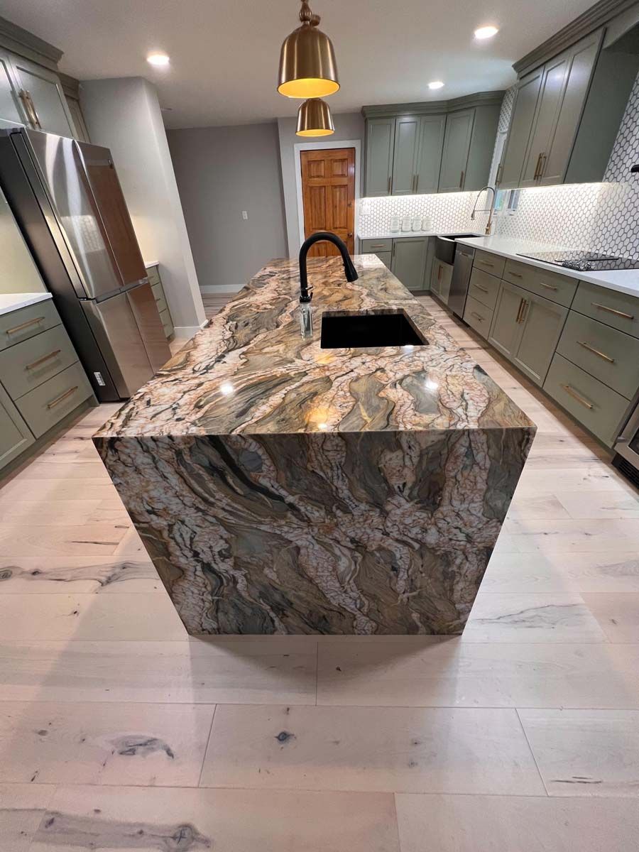 Kitchen with large marble island, black faucet, green cabinets, and light wood floors.