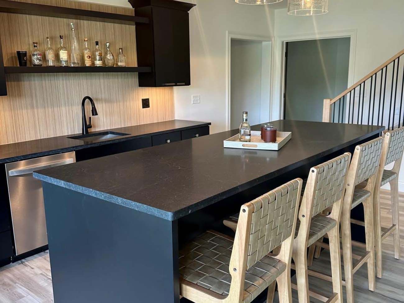 Modern kitchen with black cabinets and island, wooden bar stools, and liquor bottles on display.