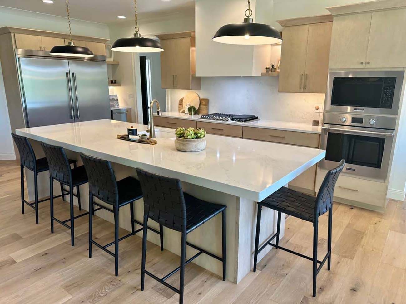 Modern kitchen with large island, black stools, light wood cabinets, and stainless steel appliances.