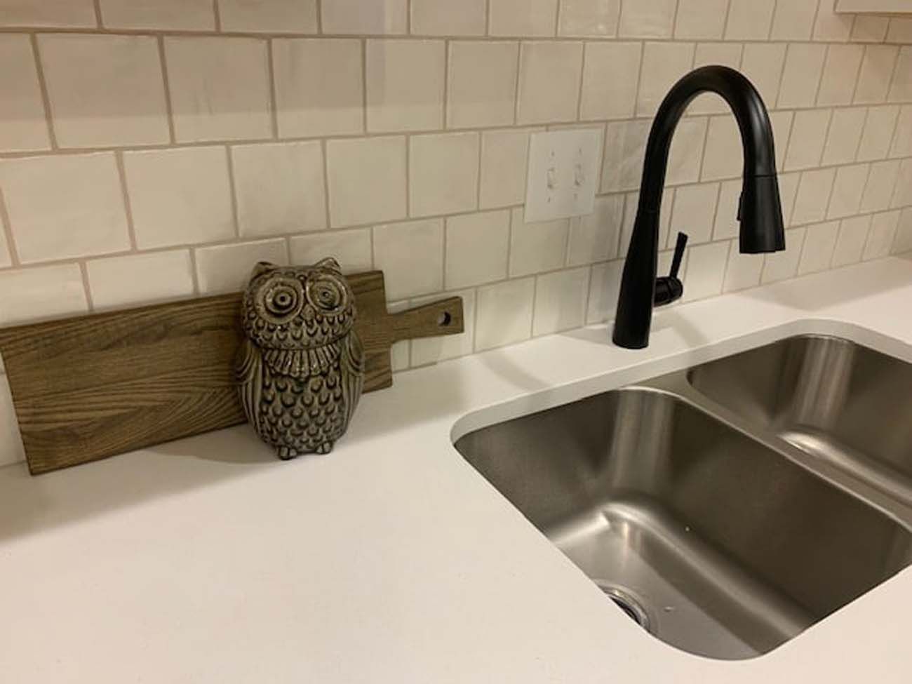 A kitchen sink with a black faucet, white countertop, and off-white tile backsplash. An owl statue and wooden cutting board sit nearby.