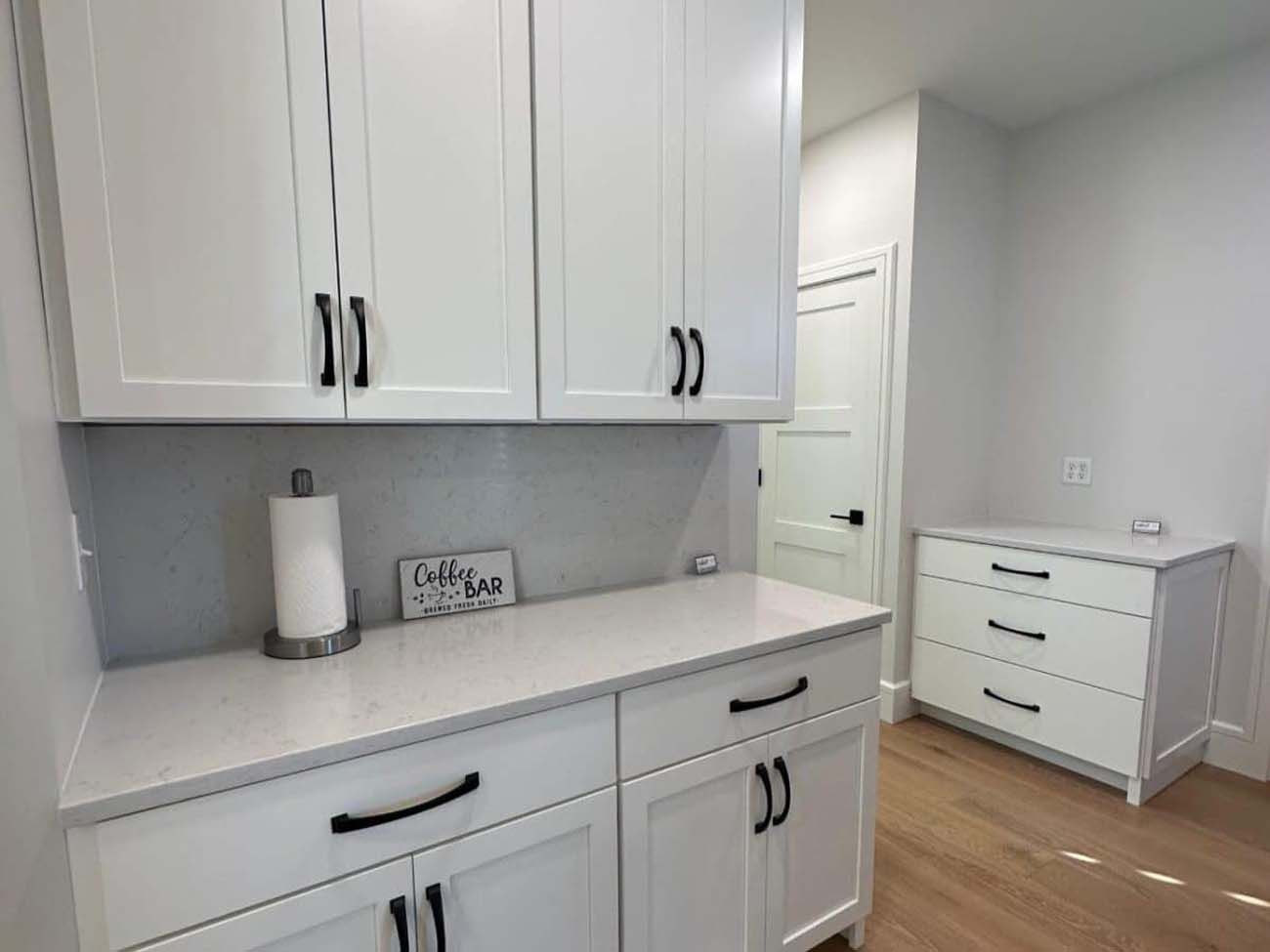 White kitchen cabinets with black hardware and a small dresser in a light-filled room.