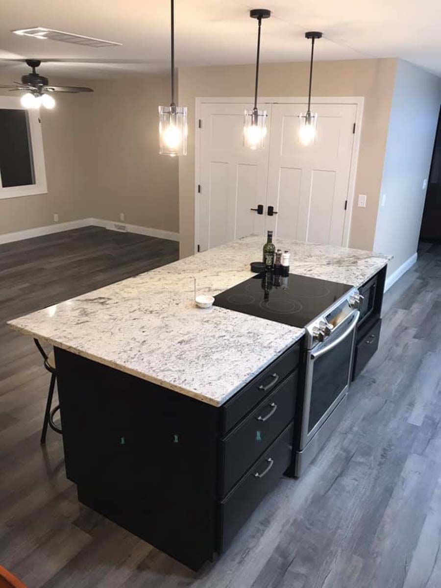 Black kitchen island with granite countertop, stovetop, oven, and drawers, under pendant lights.