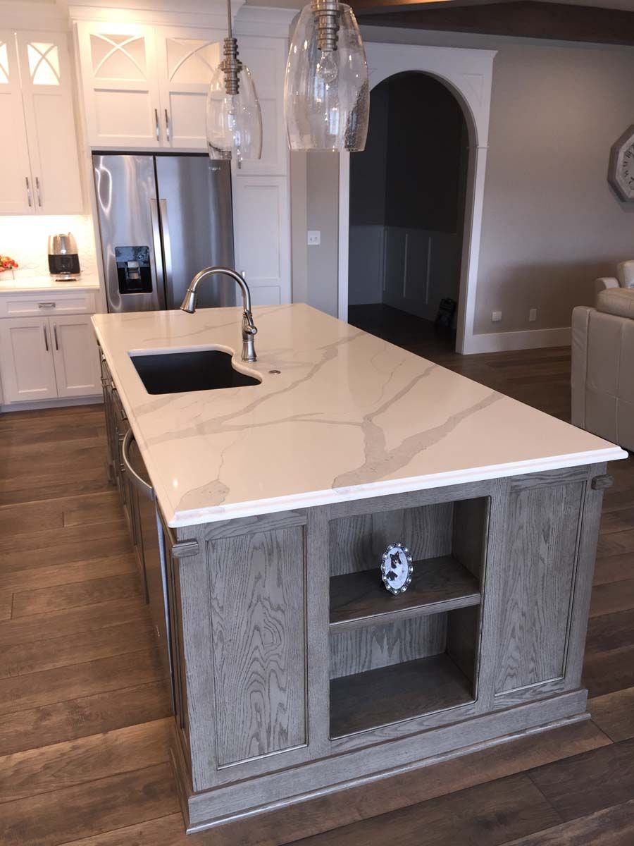 Kitchen island with a marble countertop and gray wooden cabinets, built-in sink, and shelving.